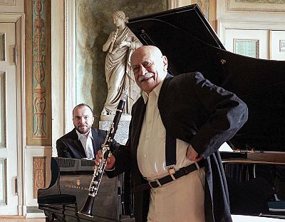 In einem barocken Konzertsaal sehen uns zwei Männer an. Einer sitzt an einem Konzertflügel und der ältere lächelt uns an, er hält eine Klarinette in der Hand.