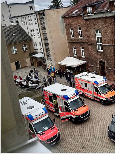 Aus einem höhergelegenen Fenster fällt der Blick in den Hof des Klinikums, in dem mehrere Rettungsfahrzeuge stehen, sowie Tische und Liegen angeordnet sind. Viele Menschen in ziviler und verschiedener Dienstkleidung sind an den Fahrzeugen und Tischen zu sehen. Sie scheinen sich in einer Pause über das Geschehen auszutauschen.