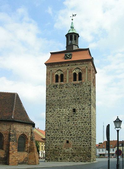 Der Marktturm steht von der Kirche getrennt. Foto: Vor uns erhebt sich ein aus Feldsteinen gemauerter Turm mit rechteckigem Grundriss. Über den Feldsteinen sind gotische Fenster in Ziegelmauerwerk gefasst, darauf sitzt ein Barockes Dach mit einer runden Turmkanzel und einer Wetterfahne. Links neben dem Turm, mit erkennbarem Abstand, ist die Apsis der St. Johanniskirche zu erkennen.